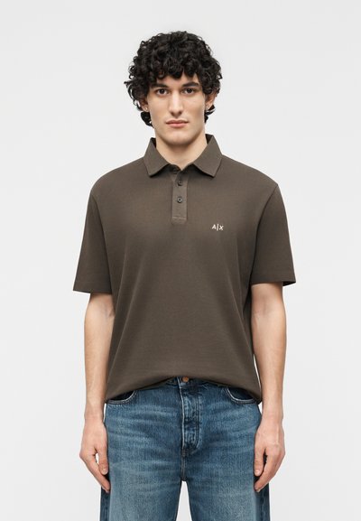 Jeune homme aux cheveux bouclés portant un polo marron à manches courtes avec trois boutons et un jean bleu, se tenant devant un fond uni.