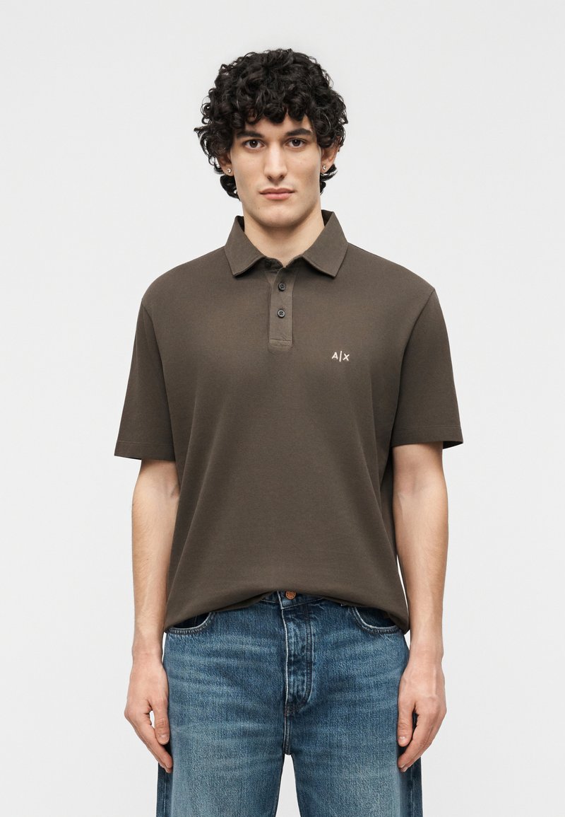 Jeune homme aux cheveux bouclés portant un polo marron à manches courtes avec trois boutons et un jean bleu, se tenant devant un fond uni.