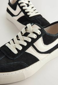 Mustat mokkakengät valkoisilla nahka-akenteilla, joissa on nauhakiinnitys ja teksturoitu kumipohja. Ompeleet reunojen ympärillä.