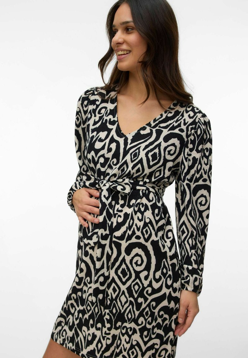 Femme souriante aux longs cheveux noirs portant une robe à manches longues à motifs noirs et blancs avec une ceinture nouée à la taille, debout devant un fond uni.