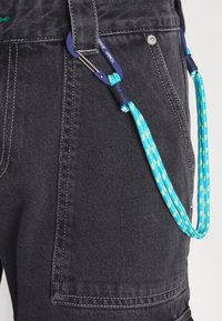 Los pantalones cargo de mezclilla negra presentan una tela texturizada con costuras reforzadas y un cordón de patrón azul y verde sujetado por un clip.