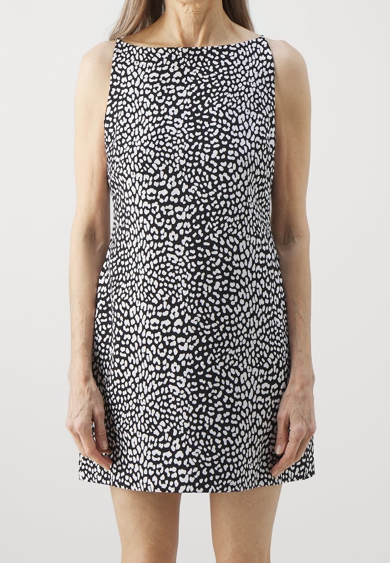 Femme portant une robe noire sans manches avec un motif abstrait à pois blancs, debout devant un fond neutre.