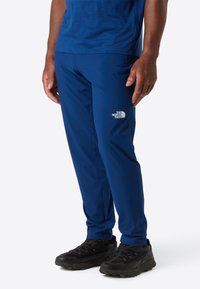 Muž v modrých kalhotách North Face a černých sportovních botách, stojící na jednobarevném pozadí.