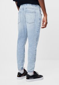 Světle modré tepláky vyrobené z denimu, s elastickým pasem a manžetami, zadními kapsami a tapered střihem. Na nohou má černé tenisky.