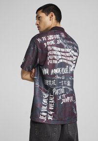 Camiseta gráfica multicolorida con patrones de texto en múltiples idiomas. Hecha de tela ligera con un corte relajado. Fondo oscuro.
