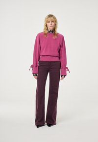 Fuchsia cropped trui met brede mouwen en strikkende details, gecombineerd met donkerv paarse flared broek. Beide kledingstukken hebben een gladde textuur.