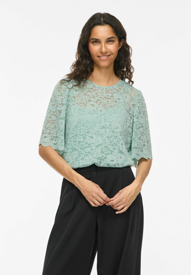 Jeune femme aux cheveux bouclés et foncés portant un chemisier en dentelle vert menthe et un pantalon large noir, souriante, les mains jointes.
