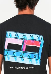 Schwarzes T-Shirt mit einem mehrfarbigen Logo in Blau, Pink und Weiß, mit dem Text "TOMMY JEANS" und Städtenamen darunter.