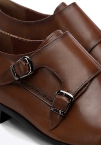 Zapatos de cuero marrón con dos hebillas decorativas, textura suave, forma redondeada y pequeñas perforaciones cerca de la zona de la hebilla.