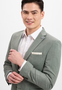 Hellgrüner Blazer aus strukturiertem Stoff, mit einem einzelnen Einstecktuch und einem weißen Hemd darunter. Der Mann passt den Ärmel des Blazers an.
