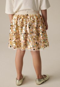 Jupe en sequins dorés et roses avec une superposition transparente, taille froncée et ceinture douce ; portée avec des ballerines en argent brillantes.