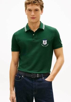 Młody mężczyzna w ciemnozielonej koszulce polo z białymi paskami na kołnierzyku i logo Tommy Hilfiger, w ciemnoniebieskich dżinsach i czarnym pasku.