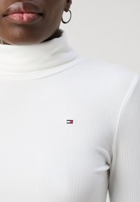 Vit ribbad polotröja med en subtil broderad logotyp i blått, rött och vitt på vänster bröstområde. Slät textur.