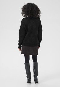 Kvinde med krøllet hår iført en sort strikket sweater, mørkebrun nederdel, sorte strømpebukser og sorte knæhøje støvler, stående med ryggen til.