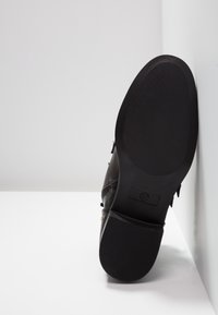 Chaussure en cuir noir avec semelle en caoutchouc texturée, dotée de détails de couture et d'accents décoratifs. Affiche le motif embossé de la semelle et le logo.