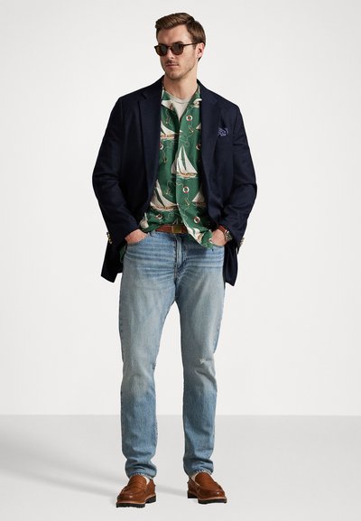 Blazer azul-marinho sobre uma camisa com padrão de barcos à vela verde, jeans azul claros e mocassins castanhos; o modelo usa óculos de sol e está de pé com as mãos nos bolsos.