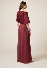 Bordeaux off-the-shoulder jumpsuit met opgeblazen mouwen, elastische tailleband, wijde pijpen en een gladde textuur. Minimalistische stijl.