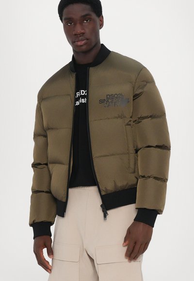 Veste matelassée vert olive avec des coutures horizontales, des accents noirs, un col haut, une fermeture éclair et un imprimé logo sur le côté supérieur gauche.