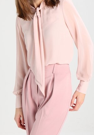 Frau trägt eine hellrosa Langarmbluse mit gebundenem Halstuch und hoch taillierte rosa Hose, stehend vor hellem Hintergrund.