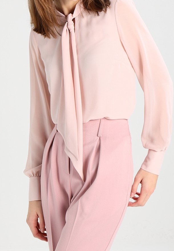 Femme portant un chemisier rose pâle à manches longues avec un foulard noué au cou et un pantalon rose taille haute, debout devant un fond clair.