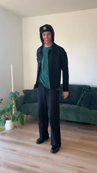 Giacca nera, camicia verde, pantaloni neri e scarpe nere. Il modello si trova in un soggiorno con un divano verde e una pianta da interno.