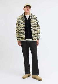 Fleece jakna u bež boji s crnim i zelenim apstraktnim uzorcima, crni kardigan, bijela košulja, tamne hlače i bež cipele s teksturiranim potplatima.