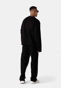 Mann mit kurzen Haaren und Sonnenbrille, der ein schwarzes Langarmoberteil und eine Hose mit weißen Turnschuhen trägt, mit dem Rücken zu einer schlichten weißen Hintergrund.