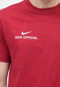 Červené bavlnené tričko s okrúhlym výstrihom a krátkymi rukávmi, na hrudi s bielym nápisom „NIKE OFFICIAL“ a logom Nike Swoosh.