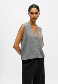 Gilet sans manches gris en tissu doux, présentant un décolleté en V profond et six boutons dorés sur le devant. Ourlet côtelé ajoutant de la texture.