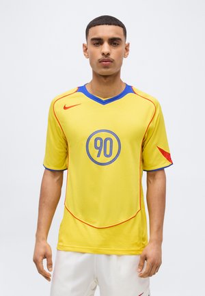 Noorikollaste servadega kollast spordisärki numbriga 90 kandva lühikeste juustega noormees, kes kannab valgeid lühikesi pükse, taustaks on ühtlane taust.