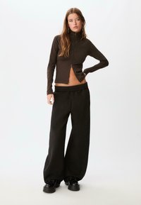Långärmad topp med hög hals i mörkbrunt tyg med en dragkedja framtill, kombinerad med vida svarta byxor med elastisk midja.