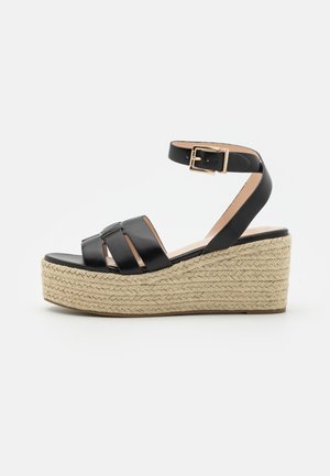 Sandale plateforme en cuir noir avec talon compensé en jute tressée et bride de cheville à boucle dorée, vue de côté sur fond blanc.