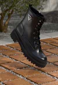 Sorte lærstøvler med spiss tå, tykk gummisåle med mønster og metalllogo-accent. Plassert mot en overflate av treverk og grus.