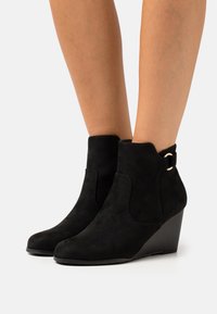Bottines en suède noir avec un talon compensé, dotées de découpes sur les côtés et d'un petit accent métallique sur la sangle de la cheville. Texture lisse.