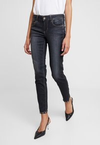 Dark denim jeans with a slim fit, medium rise, and gold accent hardware at the ankle. Paired with black stiletto heels.