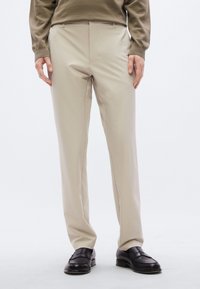 Μπεζ παντελόνι ραμμένο στα μέτρα με κομψό σχεδιασμό, λεία υφή και ελάχιστες ραφές, σε συνδυασμό με μαύρα μοκασίνια.