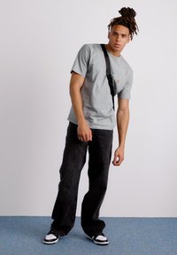 Grå kortermet t-skjorte, svarte vide bukser og svart-hvite høyhælte sneakers. Modellen bruker en crossbody veske. Glatt tekstur.