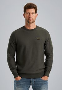 Maglione a maglia verde scuro con collo rotondo, polsini a coste e una piccola etichetta con logo sul petto. Il tessuto ha una texture liscia.