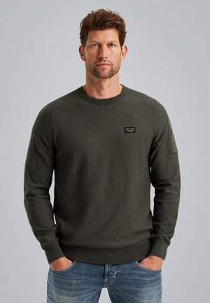 Pull en tricot vert foncé avec un col rond, des poignets côtelés et un petit patch logo sur la poitrine. Le tissu a une texture lisse.