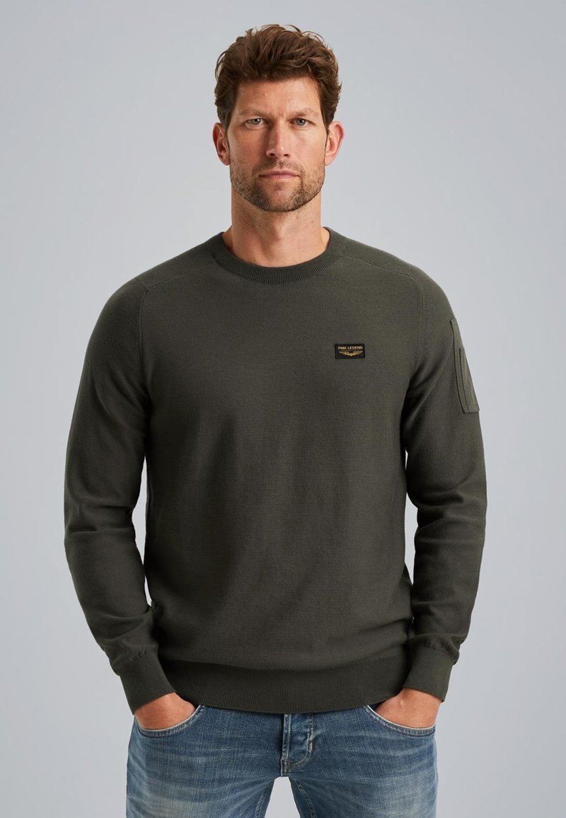 Maglione a maglia verde scuro con collo rotondo, polsini a coste e una piccola etichetta con logo sul petto. Il tessuto ha una texture liscia.