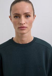Mujer joven con el cabello recogido, que lleva pendientes de aro pequeños y un sudadera verde oscuro, mirando directamente a la cámara frente a un fondo sencillo.