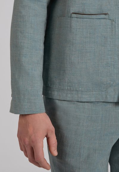 Lewa ręka obok jasnoniebieskiej marynarki lnianej oraz dopasowanych spodni z widoczną naszywaną kieszenią i obszytymi krawędziami.