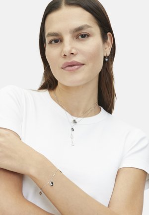 Silberne Schmuckstücke mit mehreren geschichteten Ketten, schwarz-weißen Anhängern und einem zarten Armband mit Tropfenanhängern auf einem schlichten weißen Shirt.