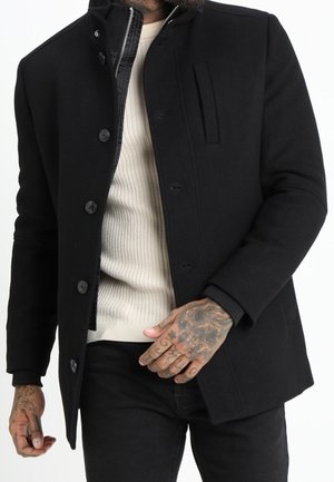 Homme portant un manteau noir boutonné par-dessus un pull beige côtelé, montrant ses mains tatouées et un pantalon noir.