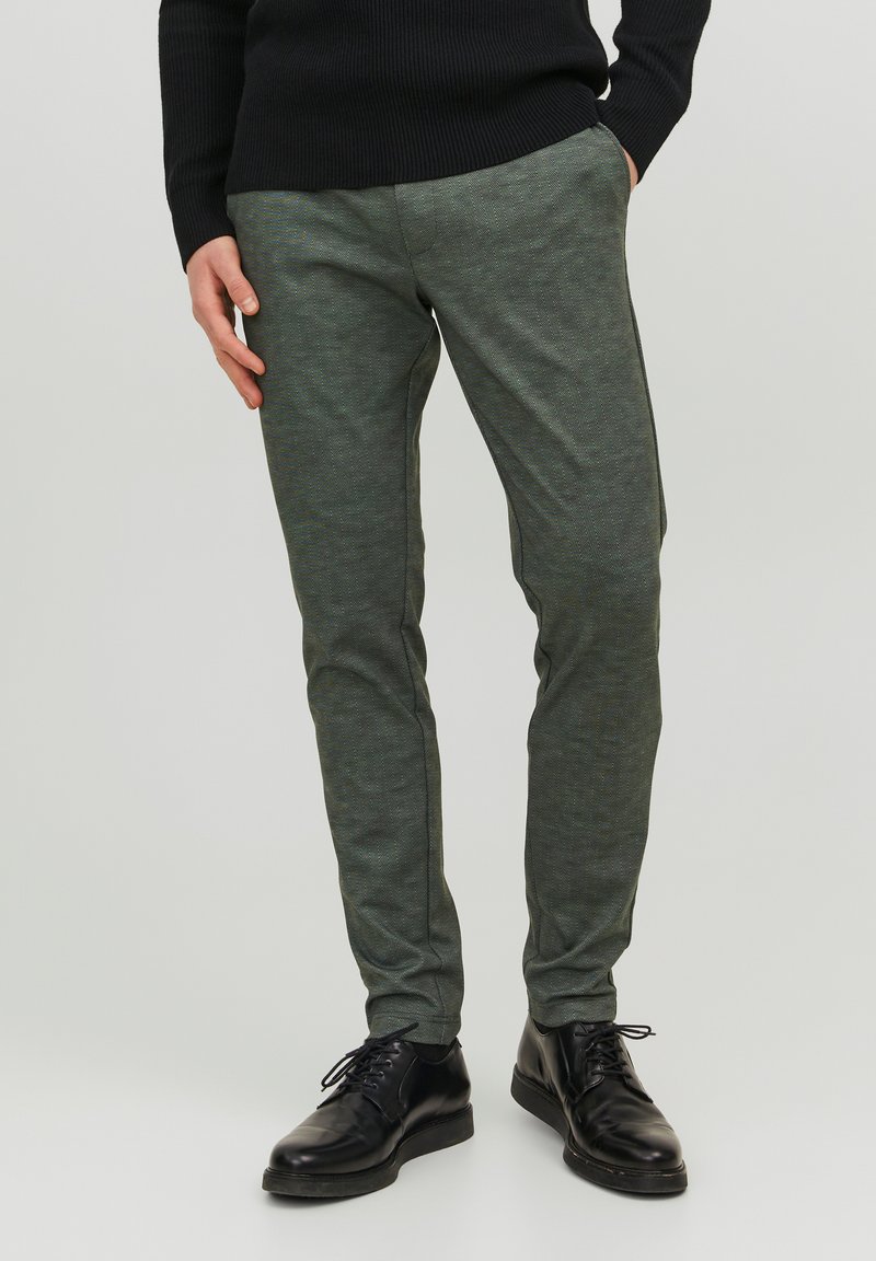 Pantaloni da uomo verde scuro realizzati in tessuto testurizzato, con una vestibilità slim e un motivo delicato, abbinati a scarpe nere stringate.