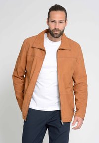 Hellbraune Lederjacke mit Reißverschluss vorne, scharfem Kragen und zwei Seitentaschen. Glatte Textur mit gesteppten Details, getragen über einem weißen Hemd.