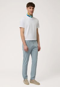 Witte kortefris shirt, lichtblauwe slim-fit broek en een kleurrijk patroon sjaal knopen om de nek. Beige instapschoenen.