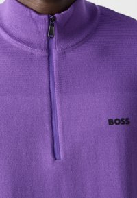 Närbild av en lila strukturerad halvdraget tröja med svart dragkedja och svart "BOSS"-logotyp på bröstet.