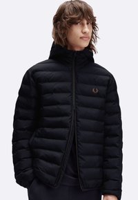 Giacca a vento nera con cappuccio, dotata di chiusura a zip e design trapuntato. Logo dorato ben visibile sulla parte sinistra del petto. Texture morbida.