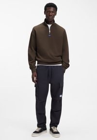 Rjava zadrga jopica, črne hlače s cargo žepi in črne superge. Gladek material, sproščen kroj, pod njo bela majica z vidnim logotipom.